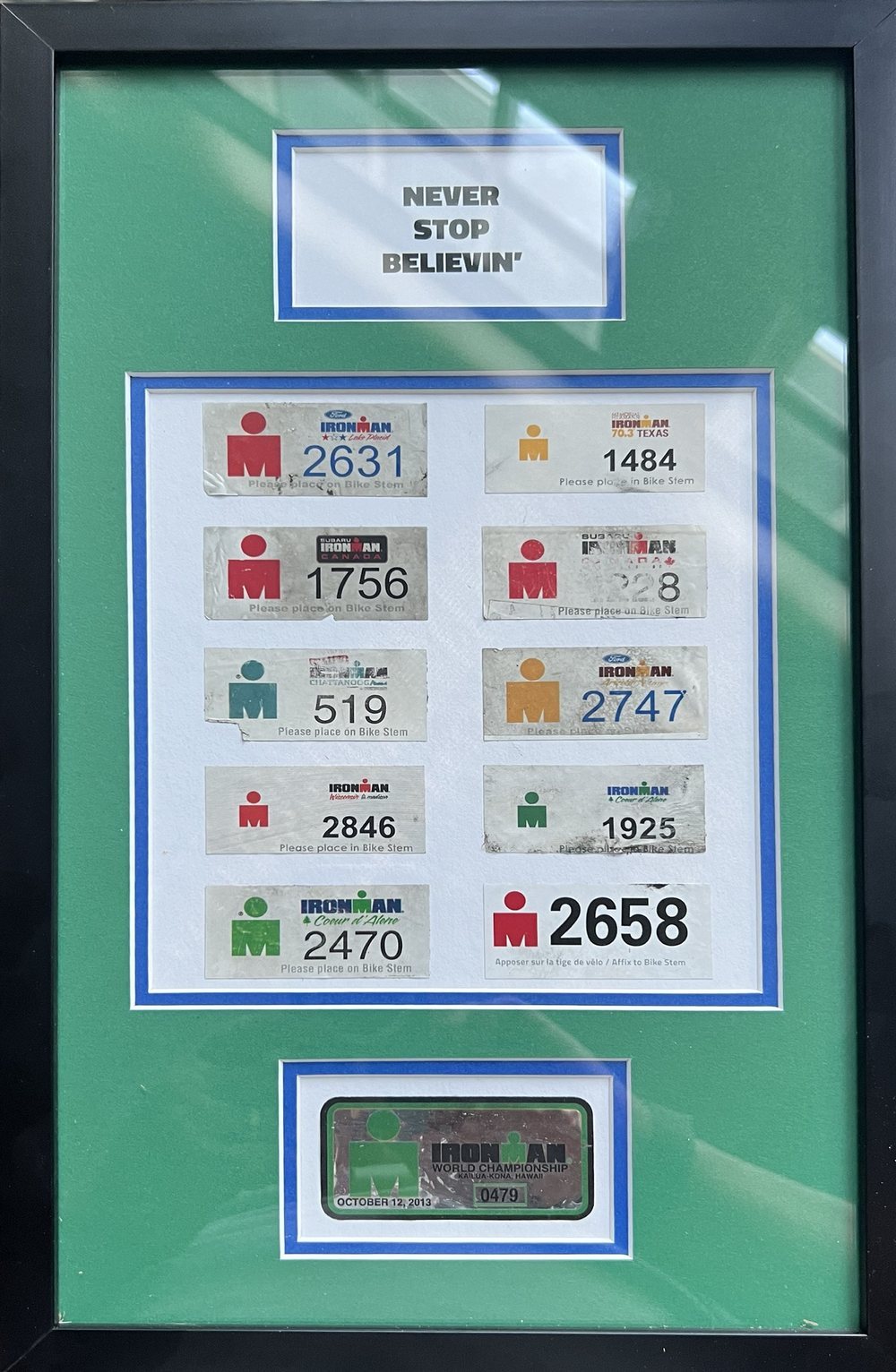 A framed set of Ironman bike-stem number plates, with the Kona World Championship plate from October 2013 beneath them